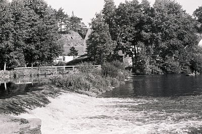 A grayscale shot features a waterway cascading over a man-made stone structure, surrounded by dense foliage & trees. A small ...