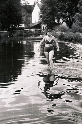 A young woman in a high-waisted, one-piece swimsuit wades through shallow water. A small house and boathouse are visible in t...