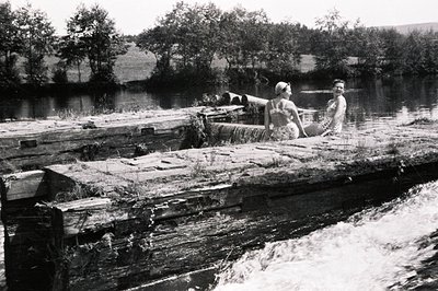 Two young women in swimwear pose on a weathered, decaying wooden structure over a river rapids. The scene captures a playful,...