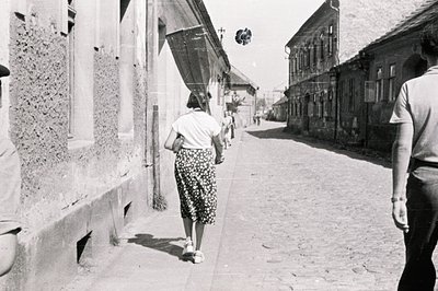 A woman in a patterned dress walks away along a cobbled street lined with aged, weathered buildings. A second figure follows ...