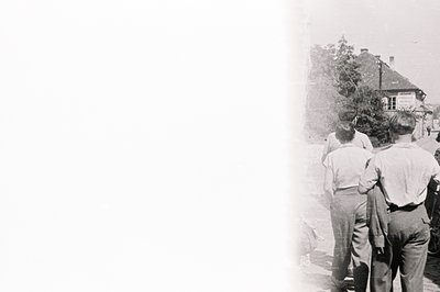 Three young men in short trousers and jackets walk away from the camera down a paved street lined with trees and a building. ...