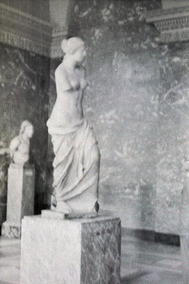 A marble statue of a draped woman stands atop a pedestal, likely within a museum or gallery. The backdrop features a patterne...