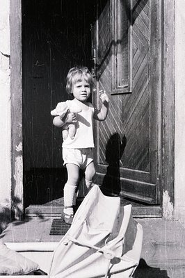 A young child, likely a toddler, stands on a doorstep clutching a teddy bear. Visible in a simple cotton dress and short pant...