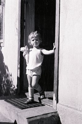 A young child stands in a doorway, holding an object (possibly bread) in their hand. The child wears a simple sleeveless top,...