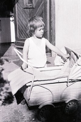 A young boy sits in a metal toy car resembling a vintage European automobile. He wears a simple sleeveless undershirt. Likely...