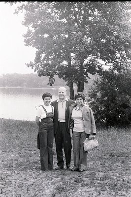 Three individuals stand lakeside, likely a family portrait. The man wears a suit jacket; the women, bell-bottom jeans and cas...