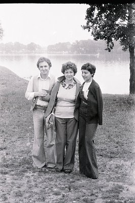 A family portrait from the 1970s: three individuals stand on a grassy bank overlooking a lake. The man wears a patterned vest...