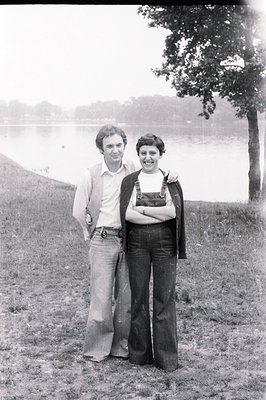 A couple stands on a grassy hill overlooking a body of water. Man in flared trousers, a button-down shirt, and a cardigan. Wo...