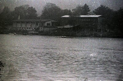 A grayscale image depicts a waterfront scene with a long, low building featuring multiple windows and a gabled roof set back ...