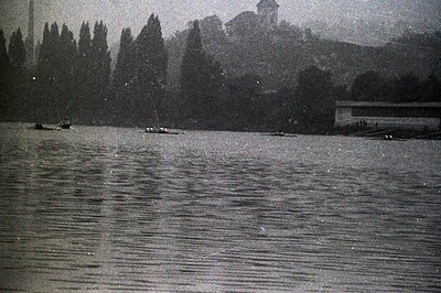 Monochrome image shows rowboats on a body of water, likely a lake or river. Trees line the shore, with a building situated on...