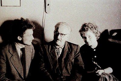 A black and white portrait depicts three individuals in a formal setting. A man with glasses sits centrally, flanked by a you...