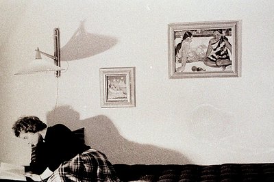 A man sits at a desk, intensely focused on papers, under the light of a wall-mounted lamp. Two framed artworks adorn the wall...