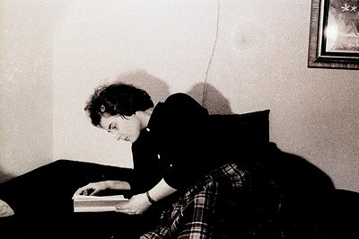 A young woman sits on a cushioned ledge, engrossed in a book. She wears a plaid shirt and black pants. A framed picture hangs...