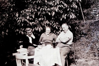 Three individuals seated at a small table covered with a white cloth outdoors. A bottle and glassware suggest a gathering. Dr...