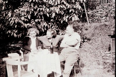 Three figures seated around a table covered with a white cloth, enjoying an outdoor meal. A wine bottle and glassware are pre...