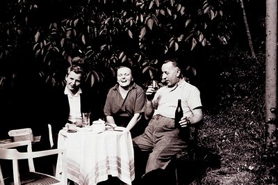 Three people seated around a table laden with drinks, captured in a candid outdoor moment. The scene, likely a garden or pati...