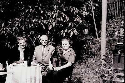 A formally dressed family—two women and a man—sit at a covered table outdoors, a small dog between them. Lush foliage fills t...