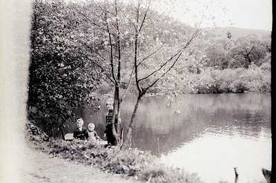 A seated woman and young child pose near a water's edge. Both wear dark, period clothing. A large tree frames the view of the...