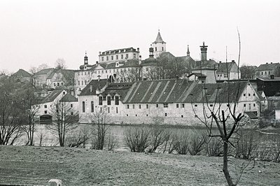 Monochrome view of a fortified structure alongside a river, likely a castle or monastery complex. Prominent white walls and g...