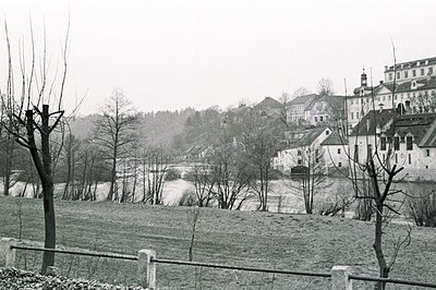 Monochromatic view of a European town, likely Central Europe, featuring a river and a large, ornate building with a steeple. ...