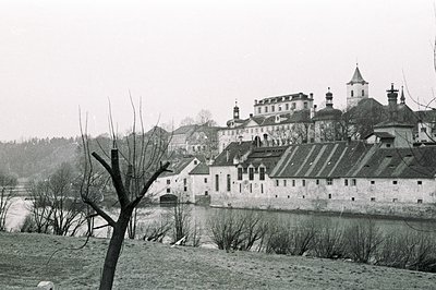 Overlooking a river, a stately castle and adjacent structures dominate the landscape. A stone wall runs parallel to the water...