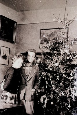 A young couple stands beside a sparsely decorated Christmas tree in a dimly lit room. The tree displays simple ornaments and ...