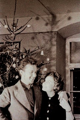 A couple embraces in front of a sparsely decorated Christmas tree, captured in a candid moment. The room features a visible b...