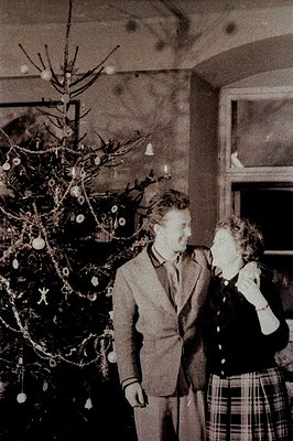 A formal portrait captures a young couple standing beside a sparsely decorated Christmas tree, likely 1950s. The man wears a ...