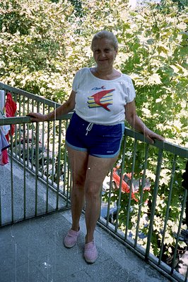 A woman stands on a balcony, hands on railing, wearing a white athletic shirt with a graphic and blue athletic shorts. Pink s...