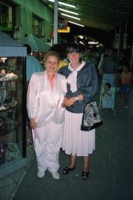 Two women stand together in what appears to be a covered market, likely a tourist area. The older woman wears a white suit wi...