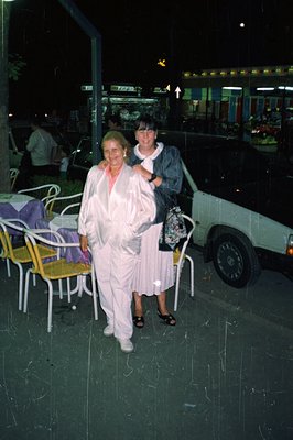 Two women in casual wear pose outdoors at night. One wears a white suit and the other, a floral dress. A restaurant patio wit...