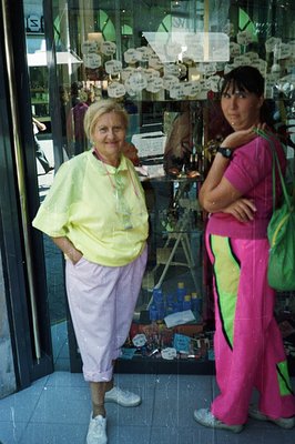 Two women stand outside a store; one in a yellow windbreaker, lilac track pants, and sneakers, the other in a pink and neon g...