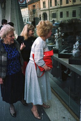 Three women examine sculptures displayed in a storefront window. The lead woman wears a blue patterned dress, the second a re...