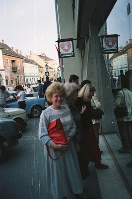 Exterior view through a shop window showing a woman in a gray dress & large hairdo holding a red purse. Visible street scene ...