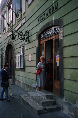 An exterior view of a building facade, displaying "RUSZWURM" signage. Two women in 1970s-era dresses stand on the entrance st...