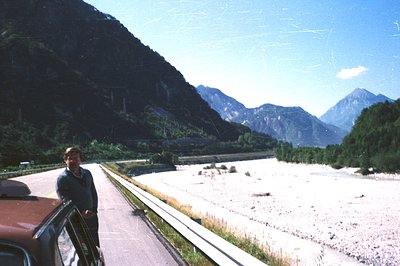 Man stands beside a vintage car on a roadside overlooking a broad, rocky riverbed framed by steep, alpine slopes and towering...