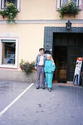 A man and woman pose outside a building with ornate window boxes and a vintage poster. The man wears a denim jacket over a sh...