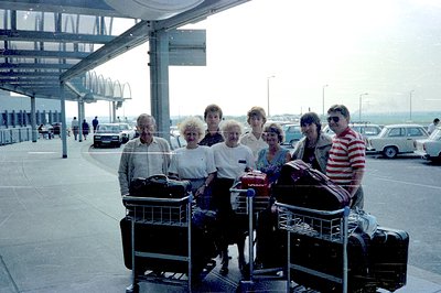 Group portrait near airport baggage claim. Six people stand with luggage carts; attire suggests a 1970s-1980s style. Cars and...