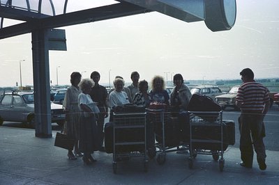 A group of travelers, likely a family, stand under a covered airport terminal. They are surrounded by stacked luggage and tro...