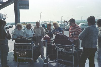 A group of vacationers, likely a family or tour group, waits at an airport curb. The scene shows luggage carts filled with ba...