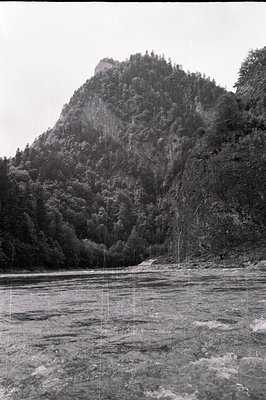 Lush, forested mountains rise dramatically from a rushing river. Dense foliage covers the steep slopes, partially obscuring r...