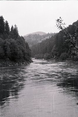 Monochrome photograph showcasing a river flowing through a deep valley. Dense evergreen forests line both banks, rising to a ...