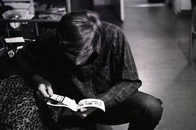 A young man, seated on a patterned sofa, intently examines a booklet or magazine. He wears a plaid shirt and dark trousers. T...