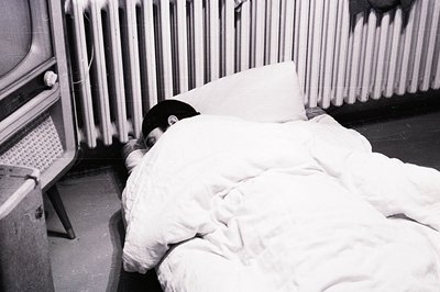 A figure lies beneath a heavy duvet, partially obscured, with only a hand visible. A vintage television set and radiator are ...