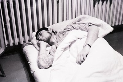 Man asleep in bed, covered with a blanket and wearing a dark cap. Visible wristwatch on his arm. Minimalist setting with a ra...