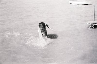 A young girl playfully splashes into shallow water. Visible details include a simple wooden dock and a portion of a boat in t...