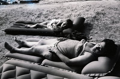 Two young women recline on inflatable mattresses in a grassy, outdoor setting, likely a backyard. They appear relaxed, some w...