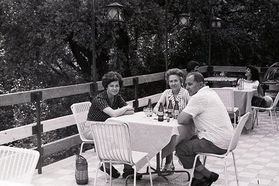 Three people sit at a table covered in a white cloth, enjoying a meal outdoors. A partial view of additional diners is visibl...