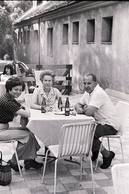 A family gathers around a table setting on a patio, likely in the 1960s. Three adults are seated – a woman in a dark floral t...