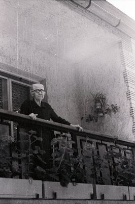 An elderly man stands on a balcony, appearing contemplative. Architectural details suggest a mid-century modern building with...